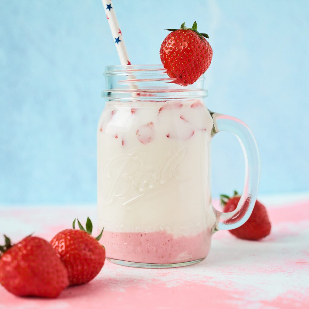 Milk with chunks of strawberry and a straw in a glass jar, surrounded by fresh strawberries.